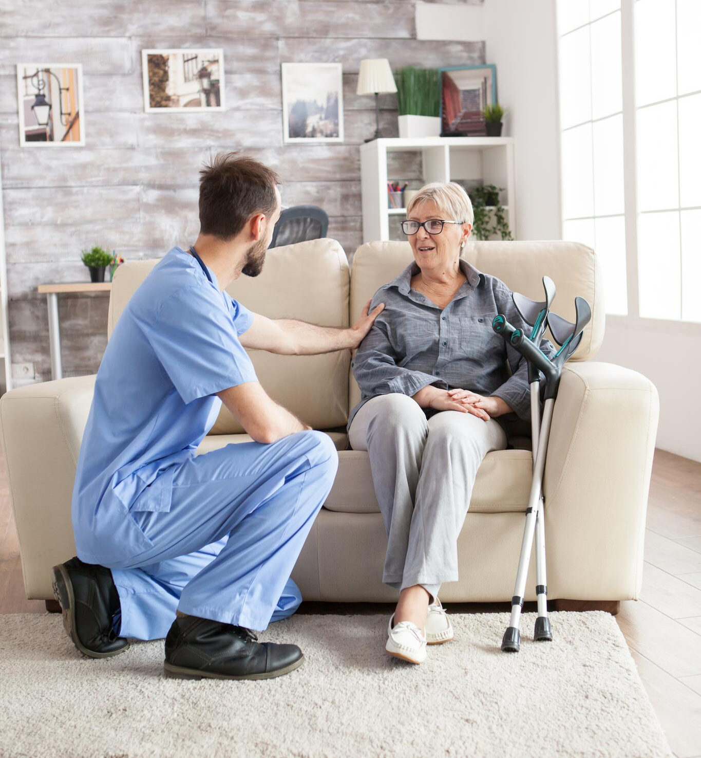 male-health-visitor-nursing-home-talking-with-retired-old-woman-while-sitting-couch-old-woman-with-crutches